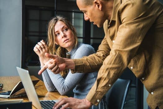 Man in Brown Long-sleeved Button-up Shirt Standing While Using Gray Laptop Computer on Brown Wooden Table Beside Woman in Gray Long-sleeved Shirt Sitting